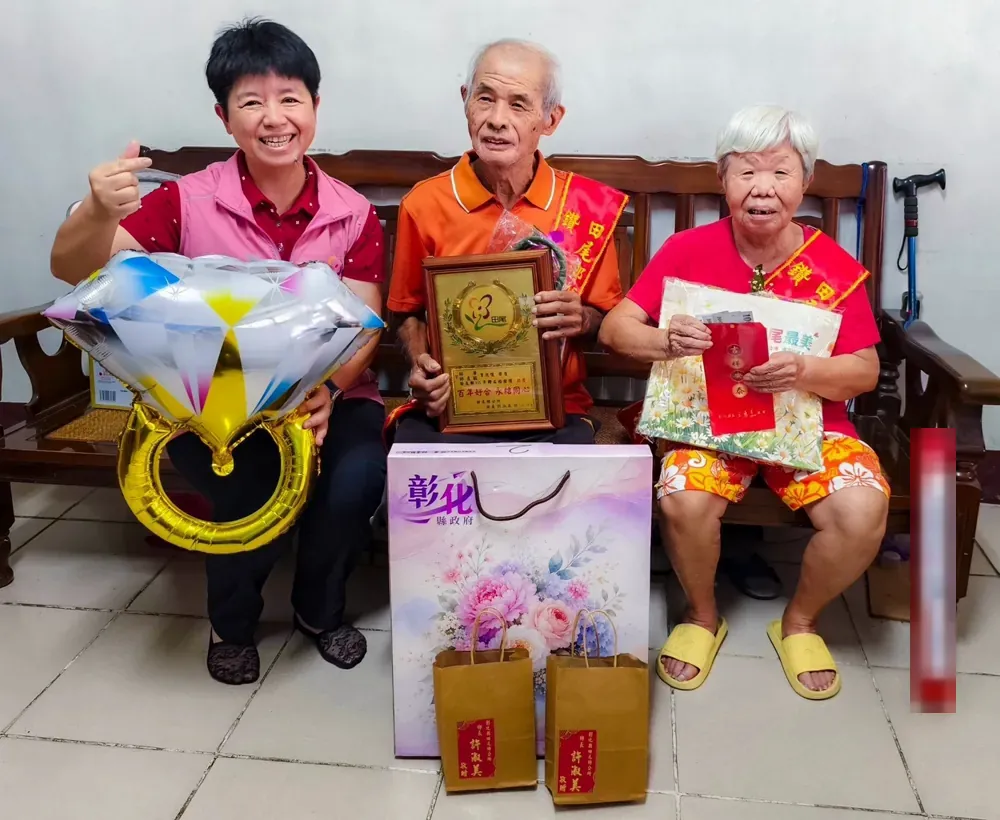 鄉長到家裡,與戴著彩帶、手捧獎牌的老夫妻合影,旁邊擺著鑽石戒指造型氣球