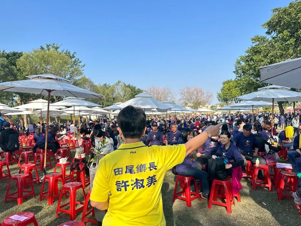 鄉長穿黃色上衣,背對鏡頭面向坐滿草地的人群揮手