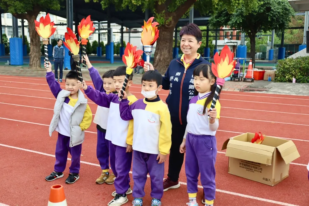 穿紫黃運動服的幼兒園孩子手拿火炬造型道具,鄉長站在旁邊